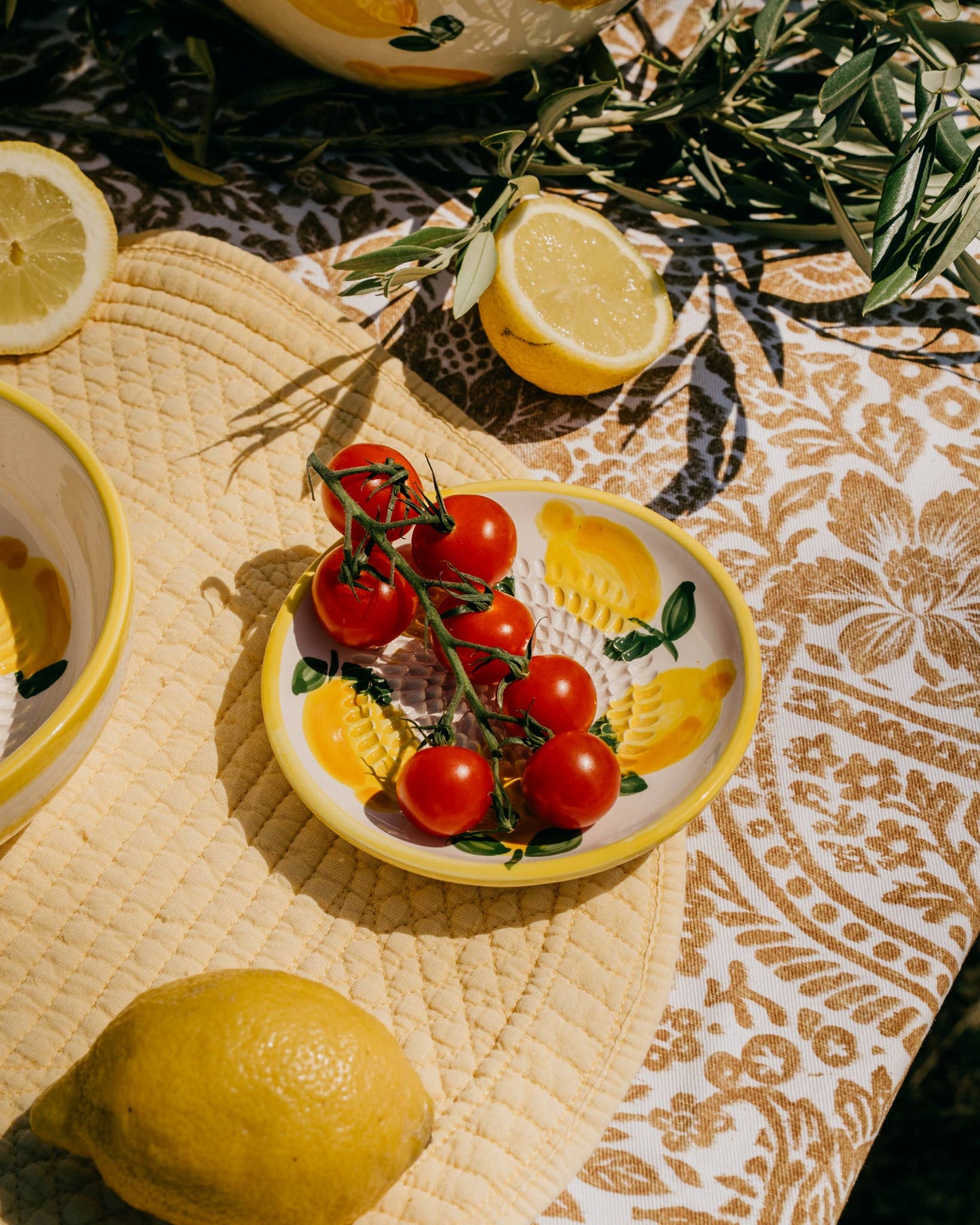 Auf dem Alfar Tierra Cocida 'Limone' Keramik Knoblauchreibe Teller (12,5 x 12,5 x 2,5 cm) liegen Kirschtomaten neben einer ganzen Zitrone, einer Zitronenhälfte und blättrigen Zweigen auf einem gemusterten Tischtuch und einem gesteppten Platzset.