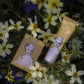 Zwei Hautpflegeprodukte - ein Stück Seife und die Sheabutter"-Handcreme von Archivist Gallery in einer goldenen Verpackung mit einem illustrierten Frauengesicht - sind inmitten blühender gelber und weißer Blumen platziert.