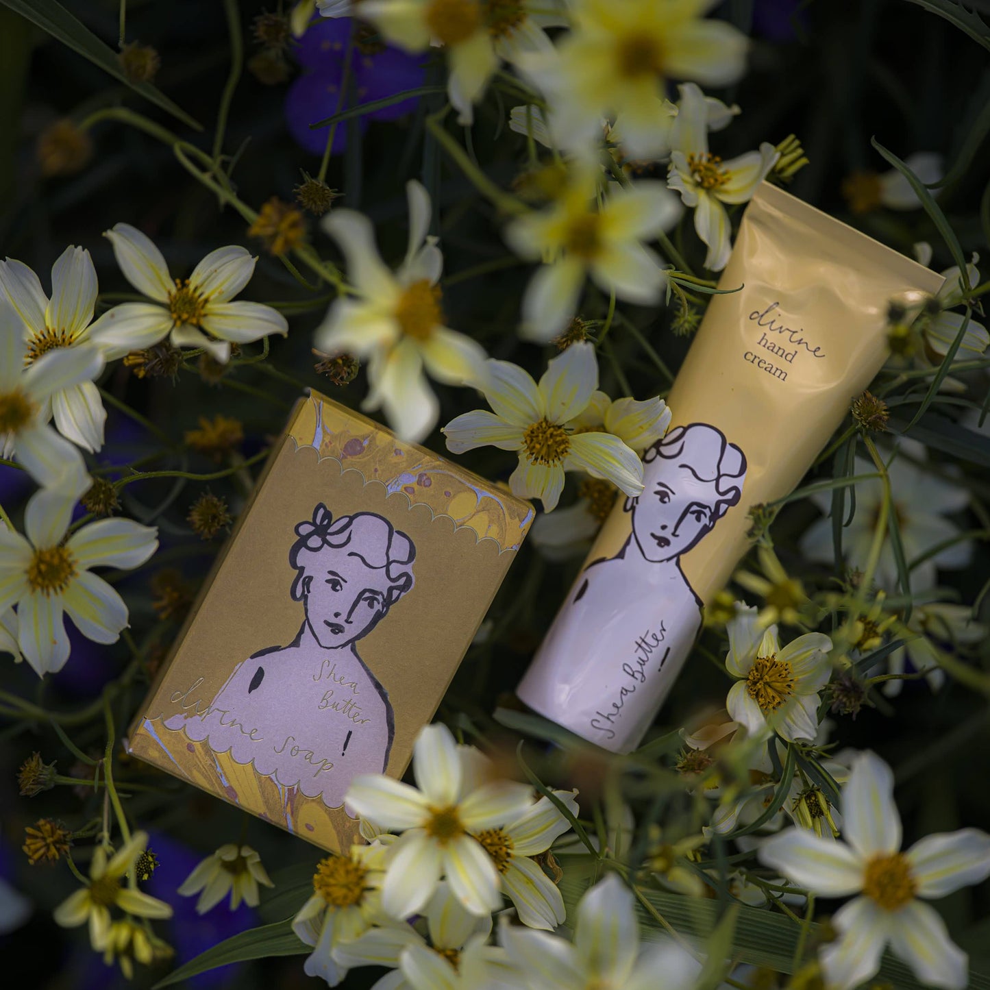 Zwei Hautpflegeprodukte - ein Stück Seife und die Sheabutter"-Handcreme von Archivist Gallery in einer goldenen Verpackung mit einem illustrierten Frauengesicht - sind inmitten blühender gelber und weißer Blumen platziert.