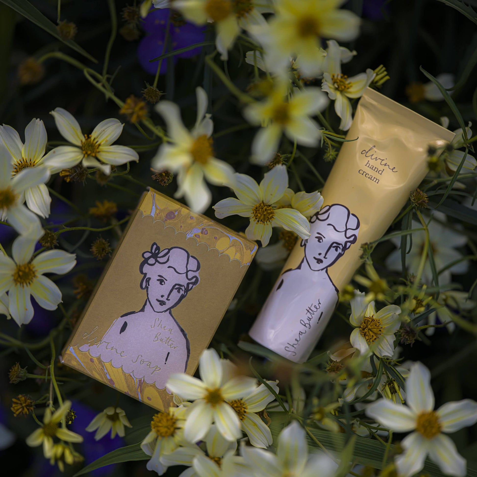 Zwei Hautpflegeprodukte - ein Stück Seife und die Sheabutter"-Handcreme von Archivist Gallery in einer goldenen Verpackung mit einem illustrierten Frauengesicht - sind inmitten blühender gelber und weißer Blumen platziert.