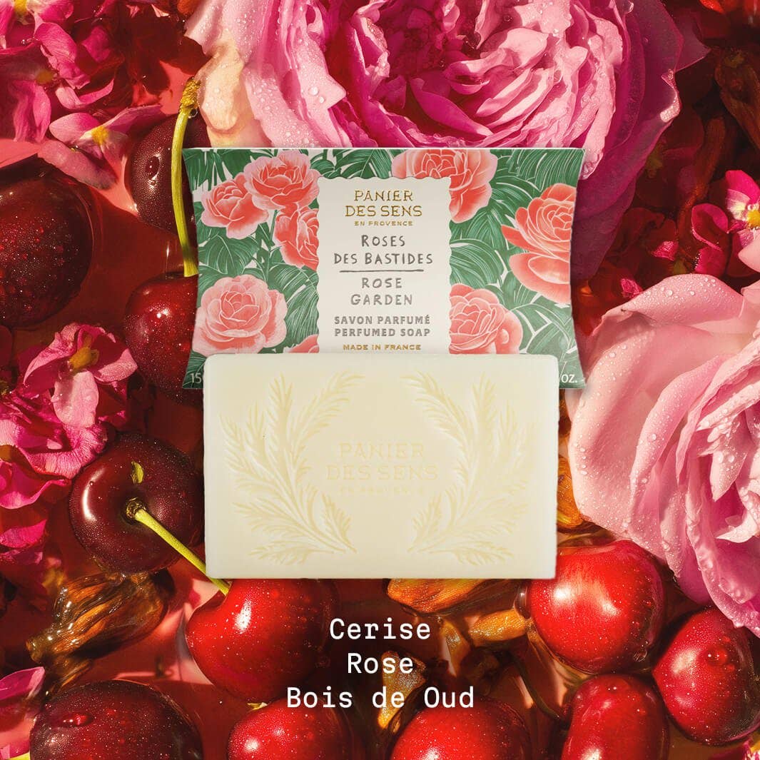 Ein Barren Panier Des Sens | Duftseife Rose des Bastides 150 g steht vor seiner blumengeschmückten Schachtel, die von rosa Rosen und Kirschen umgeben ist. Text: Cerise, Rose, Bois de Oud. Entdecke diese vegane französische Seife.