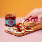 Eine Hand löffelt Firelli | Chili Crunch 200g Glas auf Cracker, die mit weißem Käse auf einem Holzbrett belegt sind; daneben stehen ein Glas Firelli, Käse und gestapelte Cracker, alles vor einem orangefarbenen Hintergrund.