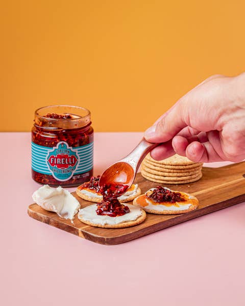 Eine Hand löffelt Firelli | Chili Crunch 200g Glas auf Cracker, die mit weißem Käse auf einem Holzbrett belegt sind; daneben stehen ein Glas Firelli, Käse und gestapelte Cracker, alles vor einem orangefarbenen Hintergrund.