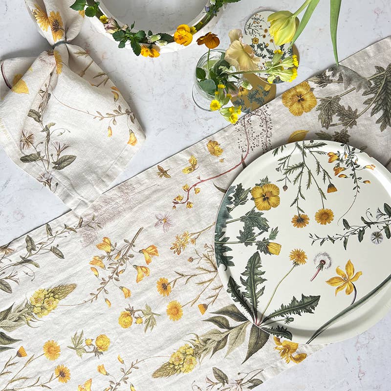 Die Tischdekoration besteht aus einem Tischläufer mit Blumenmuster, einem passenden Teller und der Koustrupco-Leinenserviette mit gelbem Blütenmotiv (45 x 45 cm). Ein Blumenkranz und eine Vase mit gelben Blüten runden dieses botanische Dekor ab.