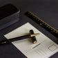Der YSTUDIO | Harz-Rollerball-Stift (Schwarz) von ENTREPÔT, ein hochwertiges Schreibgerät mit goldener Spitze, sitzt auf einer leeren Postkarte neben seinem schwarzen Etui und einem schwarz-goldenen Lineal, alles auf einer dunklen Fläche.