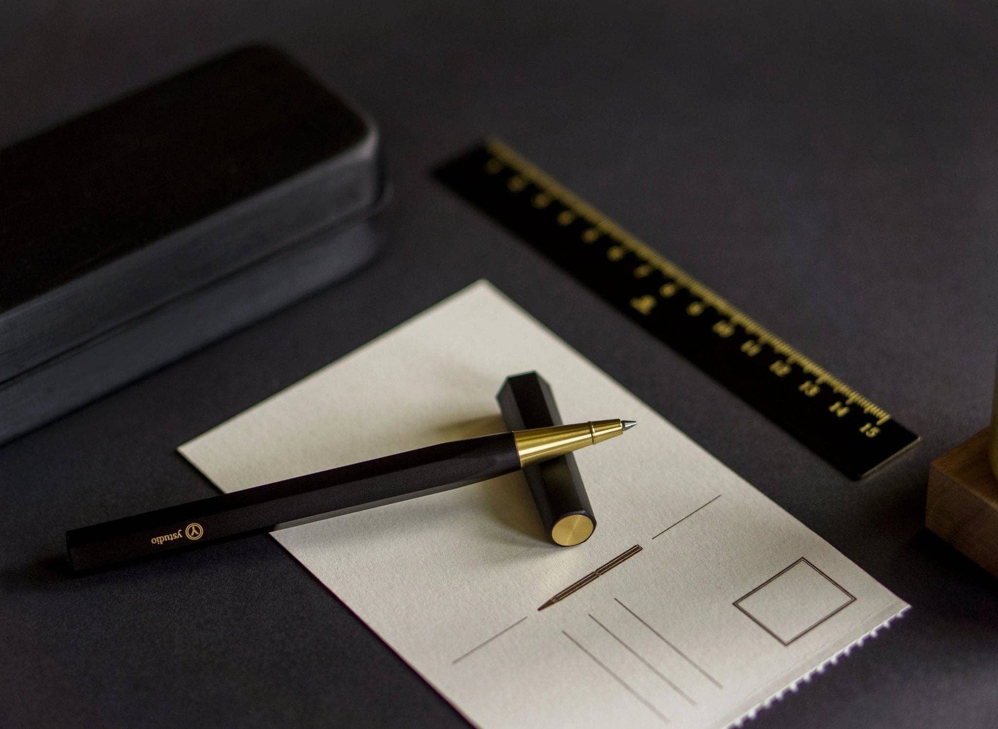 Der YSTUDIO | Harz-Rollerball-Stift (Schwarz) von ENTREPÔT, ein hochwertiges Schreibgerät mit goldener Spitze, sitzt auf einer leeren Postkarte neben seinem schwarzen Etui und einem schwarz-goldenen Lineal, alles auf einer dunklen Fläche.