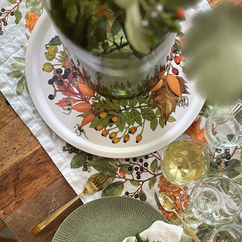 Ein elegantes Esstisch-Set besteht aus einem grünen Teller, einer goldenen Gabel, einem Weinglas und einer großen Platte mit Blumenmuster, die mit einer Vase voller frischer grüner Blätter verziert ist. Das Ganze ist wunderschön auf einem Tischläufer mit botanischem Muster arrangiert und wird durch das bezaubernde Koustrupco | Serviertablett Ø 38 cm abgerundet „Herbst“ von Koustrupco.