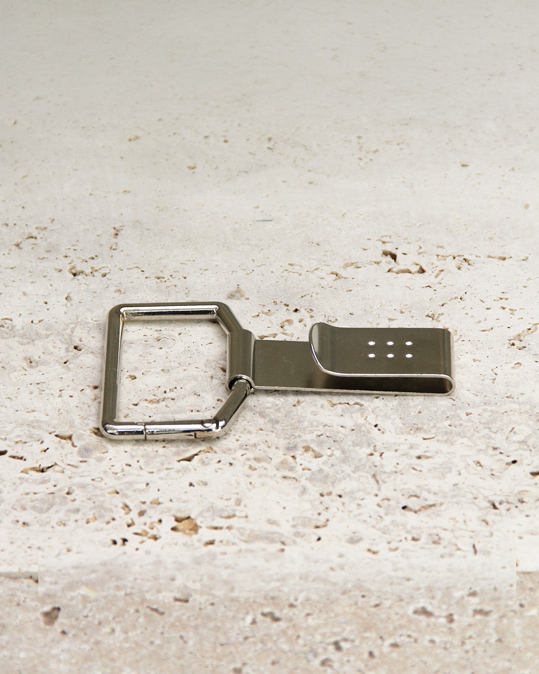 Die Niwaki 'Belt Clip' Praktische Halterung für Niwaki Single Holster, ein Metallclip mit einem rechteckigen Karabiner und kleinen quadratisch angeordneten Löchern, ruht auf strukturiertem hellem Stein - perfekt als Gartenschere Halterung oder Gürtelbefestigung.
