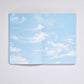 Ein aufgeschlagenes nuuna „Cloud Blue“-Inspirationsbuch in Größe M mit 176 farbigen Seiten offenbart Seiten, die mit einem blauen Himmel und zarten weißen Wolken verziert sind und das Gefühl vermitteln, in den Himmel zu blicken. Diese nuuna-Kreation liegt auf einer makellosen weißen Oberfläche und regt mit jeder farbenfrohen Seite die Kreativität an.