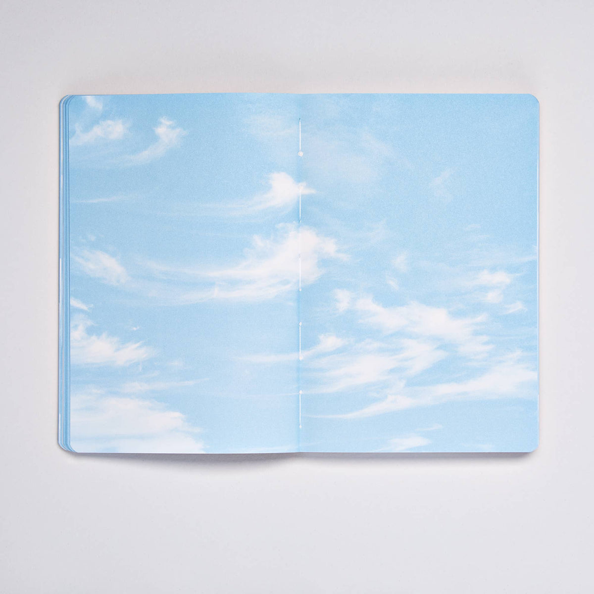 Ein aufgeschlagenes nuuna „Cloud Blue“-Inspirationsbuch in Größe M mit 176 farbigen Seiten offenbart Seiten, die mit einem blauen Himmel und zarten weißen Wolken verziert sind und das Gefühl vermitteln, in den Himmel zu blicken. Diese nuuna-Kreation liegt auf einer makellosen weißen Oberfläche und regt mit jeder farbenfrohen Seite die Kreativität an.