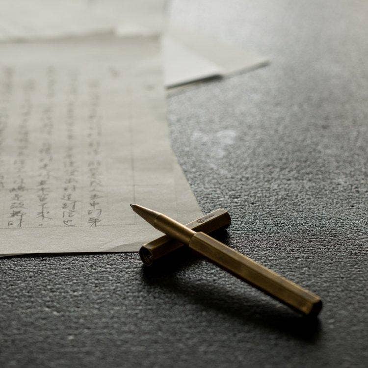 Der YSTUDIO Klassischer Revolve-Rollerball-Stift (Messing) von ENTREPÔT liegt mit seinem stabilen Griff auf einer dunklen, strukturierten Oberfläche neben Blättern mit japanischen Schriftzeichen und sorgt für eine ruhige, fokussierte Stimmung.