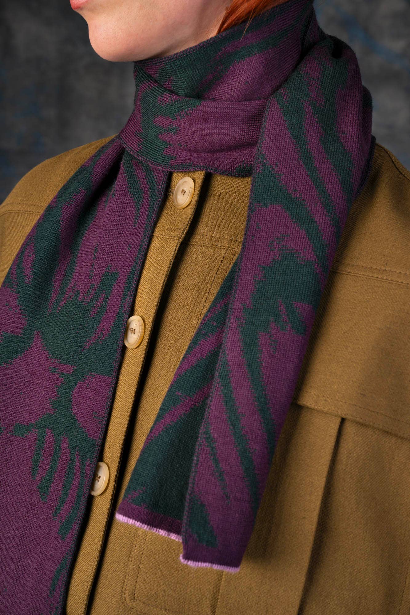 Eine Nahaufnahme zeigt eine Person in einem braunen Hemd mit Knöpfen, die den Schal aus Merinowolle - Cerura Traube Mini von Wolvis trägt, einen wendbaren Jacquard-Schal mit dunkelgrünen Mustern. Nur die untere Hälfte des Gesichts ist teilweise sichtbar.