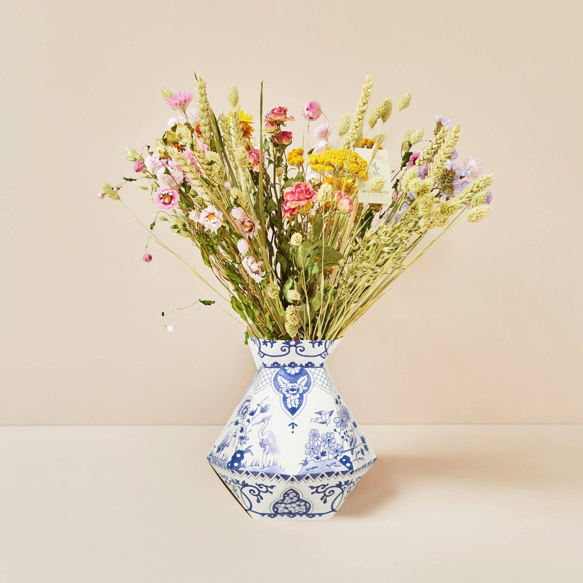 In einer blau-weißen Ziervase steht ein Strauß aus Trockengräsern und Wildblumen aus der Kollektion „Feldblumenstrauß“ S – M von Wildflowers by Floriette, der vor einem schlichten beigen Hintergrund präsentiert wird.
