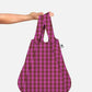 Eine Hand hält die Notabag | Wandelbare Tasche und Rucksack „Kirsche“ von Notabag in der Hand, wobei das rosa-braune Karomuster vor einem schlichten weißen Hintergrund zur Geltung kommt und das clevere Design hervorgehoben wird, dank dem sich die Tasche mühelos von einer Tragetasche in einen stilvollen Rucksack verwandeln lässt.
