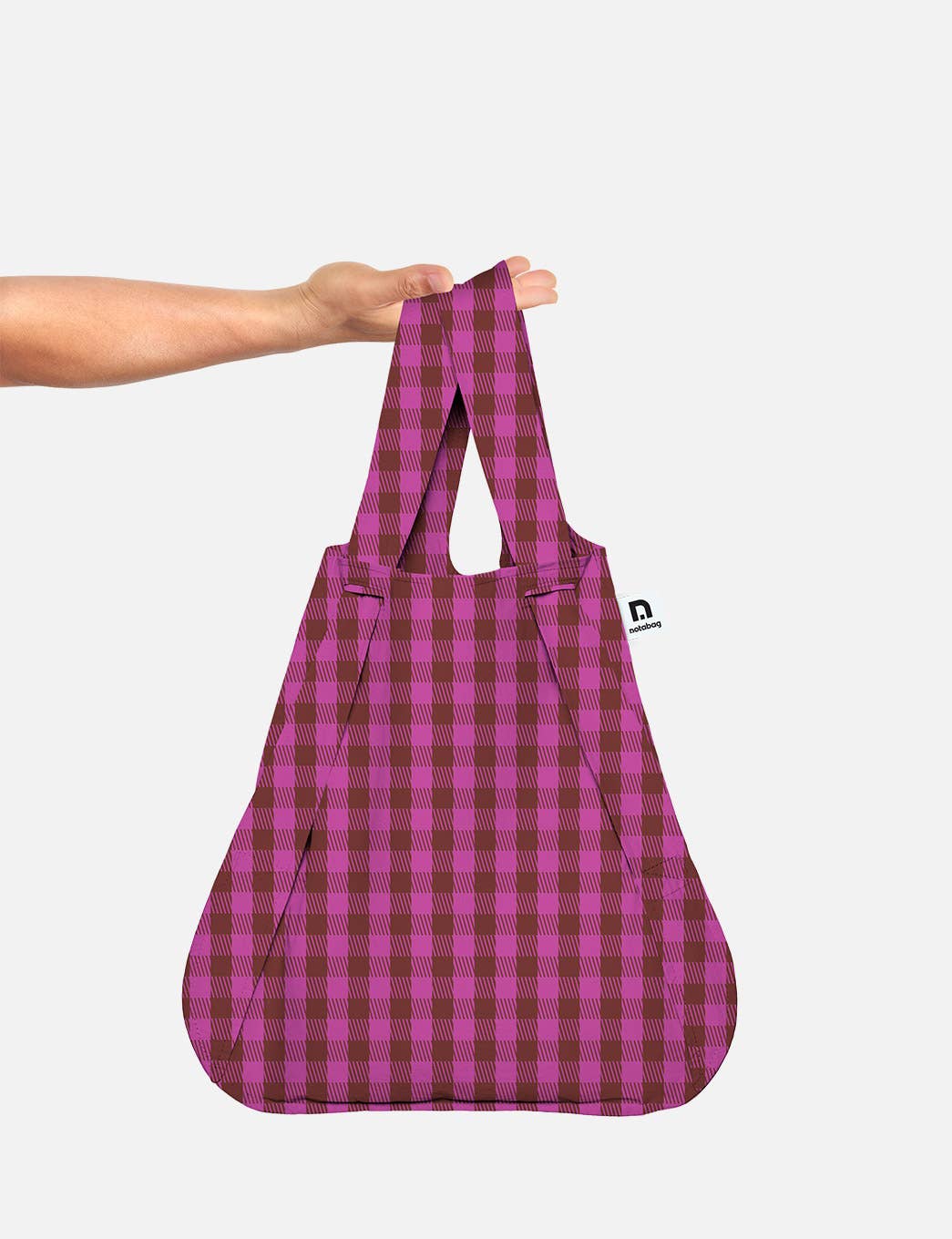 Eine Hand hält die Notabag | Wandelbare Tasche und Rucksack „Kirsche“ von Notabag in der Hand, wobei das rosa-braune Karomuster vor einem schlichten weißen Hintergrund zur Geltung kommt und das clevere Design hervorgehoben wird, dank dem sich die Tasche mühelos von einer Tragetasche in einen stilvollen Rucksack verwandeln lässt.