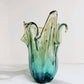 Die Murano Dream | Die mittelgroße „Giusy“-Vase aus grünem Murano-Glas (H 24, Ø 18 cm) besticht durch ihr handgefertigtes, wellenförmiges Design mit skulpturalem Rand und kommt auf weißem Marmor vor hellem Hintergrund besonders elegant zur Geltung.
