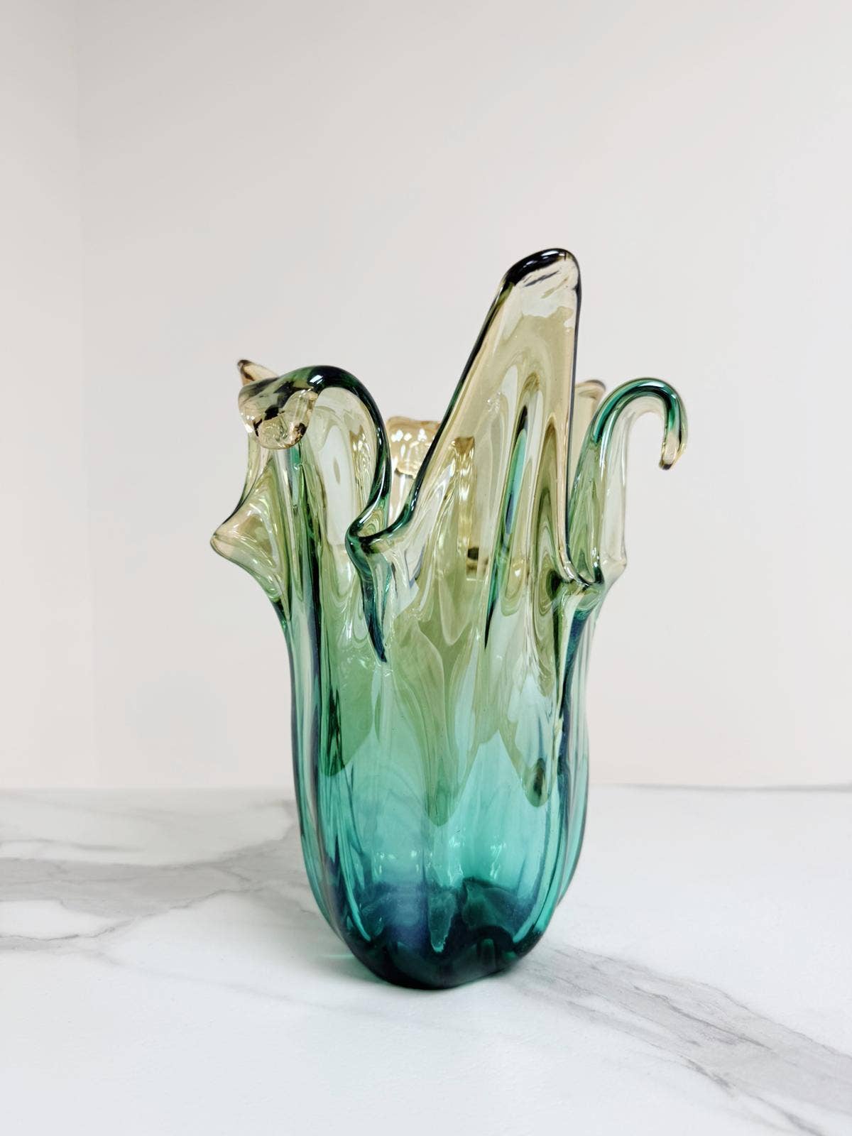 Die Murano Dream | Die mittelgroße „Giusy“-Vase aus grünem Murano-Glas (H 24, Ø 18 cm) besticht durch ihr handgefertigtes, wellenförmiges Design mit skulpturalem Rand und kommt auf weißem Marmor vor hellem Hintergrund besonders elegant zur Geltung.