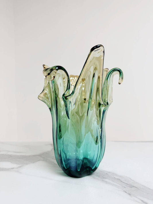 Die Murano Dream | Die mittelgroße „Giusy“-Vase aus grünem Murano-Glas (H 24, Ø 18 cm) besticht durch ihr handgefertigtes, wellenförmiges Design mit skulpturalem Rand und kommt auf weißem Marmor vor hellem Hintergrund besonders elegant zur Geltung.