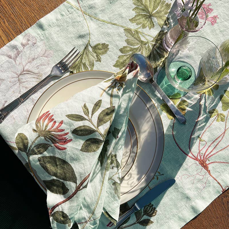 Die Tischdekoration besteht aus dem Leinen-Tischläufer „Flora Danica Atlas“ von Koustrupco (145 x 45 cm) mit grünen Blättern und rosa Blüten, ergänzt durch Teller mit Blumenmuster, eine dazu passende Serviette, Besteck und ein Glas mit grünem Fuß, das im Sonnenlicht schimmert.