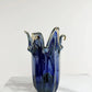 Die „Murano Dream Große Giusy“-Vase aus blauem Original-Murano-Glas (H 32, Ø 20 cm) steht auf weißem Marmor; ihre skulpturale Form mit wellenförmigen Rändern und die Akzente aus klarem Glas kommen vor dem schlichten, hellen Hintergrund besonders gut zur Geltung.