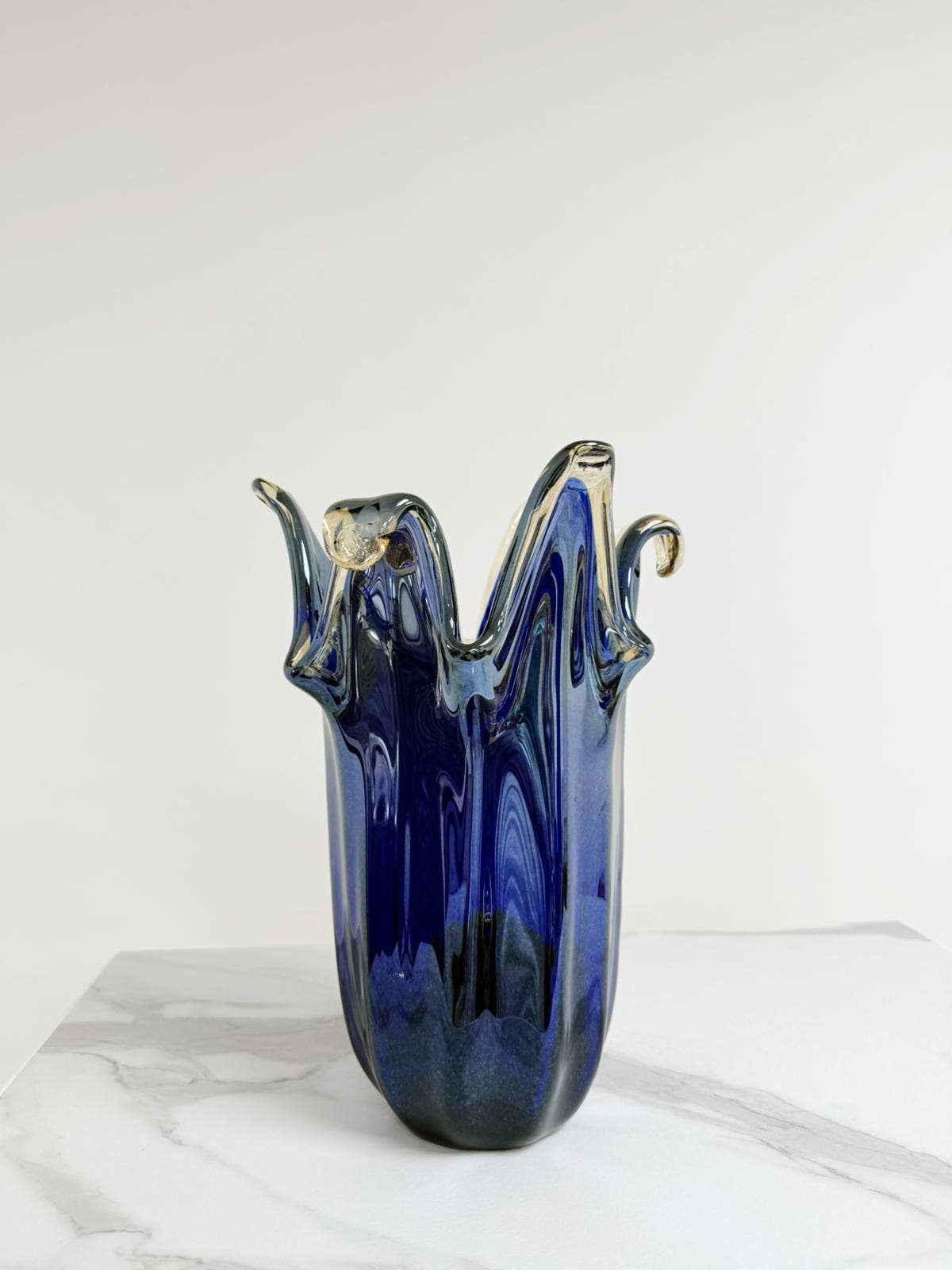 Die „Murano Dream Große Giusy“-Vase aus blauem Original-Murano-Glas (H 32, Ø 20 cm) steht auf weißem Marmor; ihre skulpturale Form mit wellenförmigen Rändern und die Akzente aus klarem Glas kommen vor dem schlichten, hellen Hintergrund besonders gut zur Geltung.