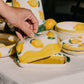 Eine Hand hebt den Deckel der Alfar Tierra Cocida | Handgefertigte Keramik-Butterdose „Limone“ in Gelb und Weiß an – eine mit gelben Zitronen verzierte Keramik-Butterdose von Alfar Tierra Cocida, umgeben von passender handbemalter Keramik und anderen mediterranen Tischartikeln auf einem wunderschön gedeckten Tisch.