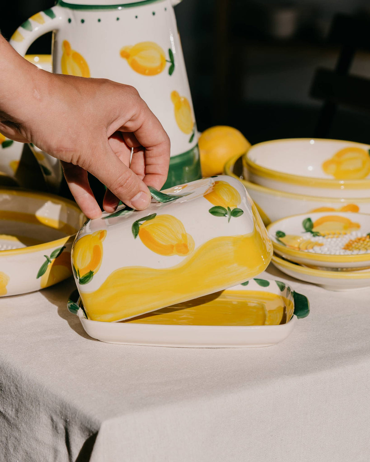 Eine Hand hebt den Deckel der Alfar Tierra Cocida | Handgefertigte Keramik-Butterdose „Limone“ in Gelb und Weiß an – eine mit gelben Zitronen verzierte Keramik-Butterdose von Alfar Tierra Cocida, umgeben von passender handbemalter Keramik und anderen mediterranen Tischartikeln auf einem wunderschön gedeckten Tisch.