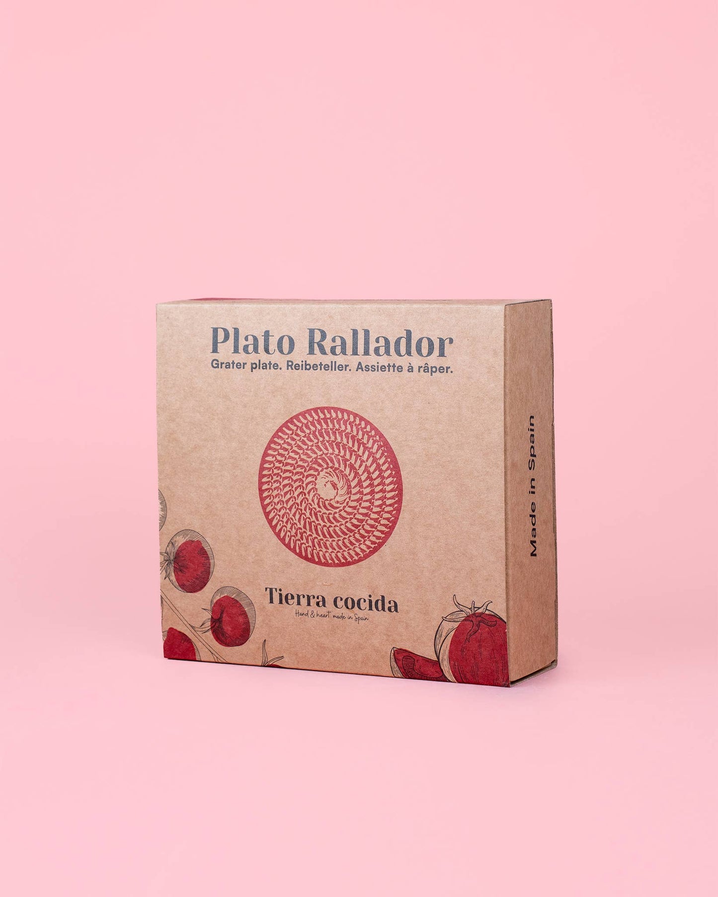 Eine braune Pappschachtel mit der Aufschrift Alfar Tierra Cocida zeigt ein rotes Kreismuster, Tomaten- und Zitronenmotive und deutet die Keramik Knoblauchreibe "Limone" im Inneren an. "Tierra cocida" und "Made in Spain" sind auf der Seite aufgedruckt, vor einem rosa Hintergrund.