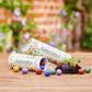 Zwei Röhrchen mit der Aufschrift Stadtgärtner | 25 bunte Blumenmurmeln in Dose liegen auf einer Fläche. Bunte Blumensamenbomben quellen hervor, einige sprießen als kleine Pflanzen in Bodennähe. Ein verschwommener Garten dient als Hintergrund. Marke: Stadtgärtner.