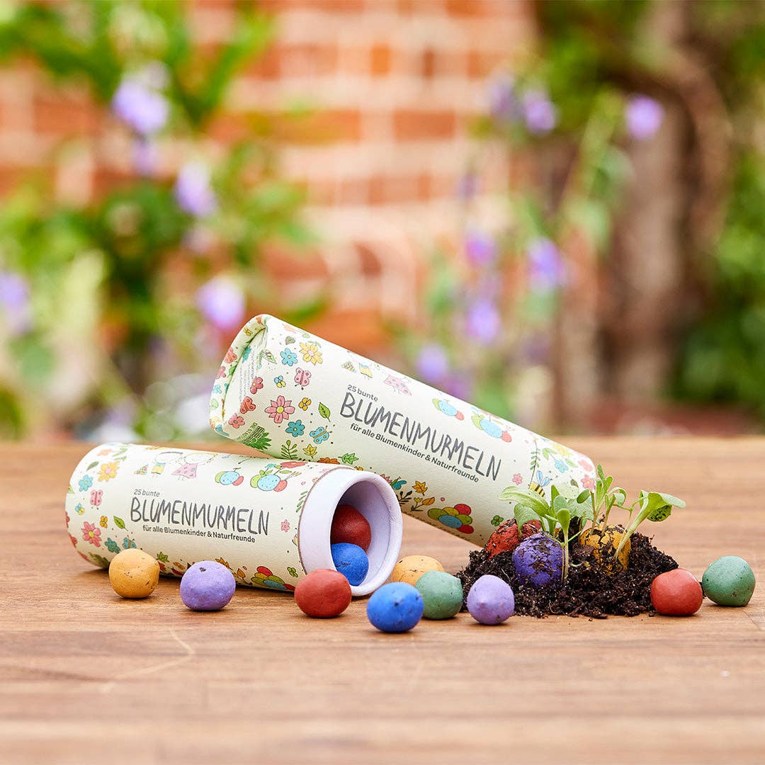 Zwei Röhrchen mit der Aufschrift Stadtgärtner | 25 bunte Blumenmurmeln in Dose liegen auf einer Fläche. Bunte Blumensamenbomben quellen hervor, einige sprießen als kleine Pflanzen in Bodennähe. Ein verschwommener Garten dient als Hintergrund. Marke: Stadtgärtner.