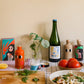 Eine Küchenszene mit bunten Flaschen, Tomaten, Nudeln, einer Zitronenspalte, Kräutern und Yiayia and friends Natives Olivenöl extra mit Chili auf einem Holzbrett und kariertem Tuch erweckt die mediterrane Würze vor einer beigen Wand zum Leben.