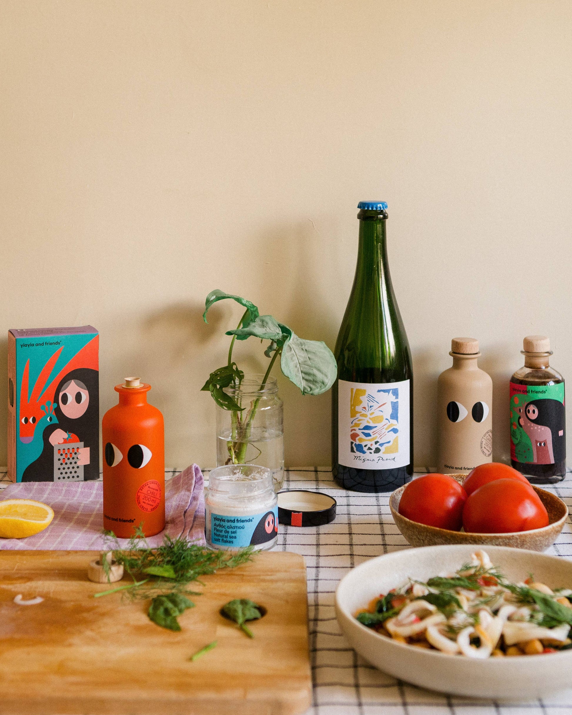 Eine Küchenszene mit bunten Flaschen, Tomaten, Nudeln, einer Zitronenspalte, Kräutern und Yiayia and friends Natives Olivenöl extra mit Chili auf einem Holzbrett und kariertem Tuch erweckt die mediterrane Würze vor einer beigen Wand zum Leben.