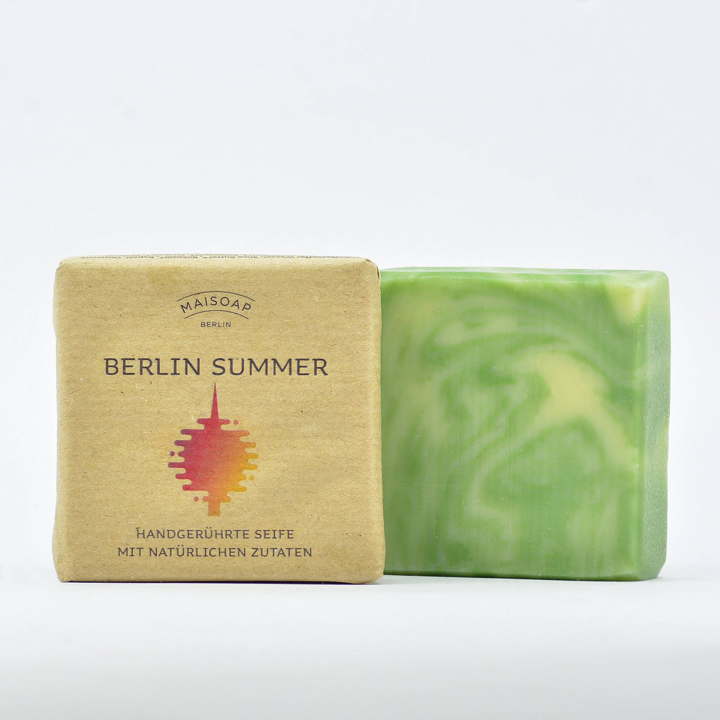 Zwei Riegel Maisoap | Berlin Summer Seife von Maisoap: einer in einer braunen Papierverpackung mit roter und gelber Grafik, der andere ausgepackt, um ein grün-weiß marmoriertes Design zu zeigen, beide präsentiert auf einem schlichten weißen Hintergrund.