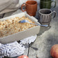 Eine Auflaufform mit goldenen Streuseln und einem Löffel steht auf einem rustikalen Tisch mit Äpfeln, einem gemusterten Tuch und zwei Sagaform | Kaffeebecher terrakotta, 2-teilig von Sagaform GmbH. Das Sonnenlicht wirft sanfte Schatten auf die Szene.