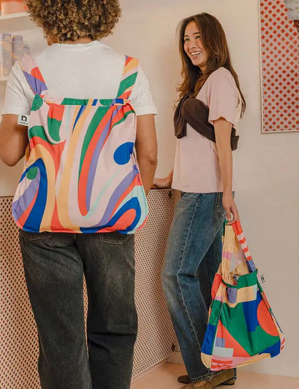 Zwei Personen mit bunter Notabag | 'Flow' Kombination aus Tasche & Rucksack von Notabag stehen in einem hellen Raum. Die eine trägt die Tasche als Rucksack, die andere trägt lächelnd die umweltfreundliche Tasche an ihrer Seite.