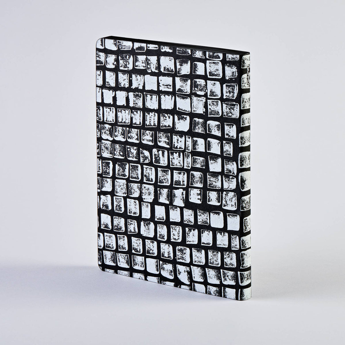 Ein geschlossenes nuuna | „Sassy Squares“ Notizbuch Not Ordinary L Light A5+ von nuuna mit einem Einband in schwarz-weißem Karomuster steht aufrecht auf einer schlichten, hellen Fläche.