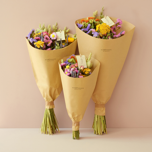Drei „Feldblumenstrauß“-Blumensträuße in den Größen S–M aus der Kollektion „Wildflowers by Floriette | Trockenblumen“ sind in braunes Papier eingewickelt und vor einem hellrosa Hintergrund präsentiert. Jeder Blumenstrauß ist mit einem Anhängeschild versehen und besticht durch eine farbenfrohe Mischung aus gelben, violetten und rosa Trockenblumen mit sichtbaren grünen Stielen.