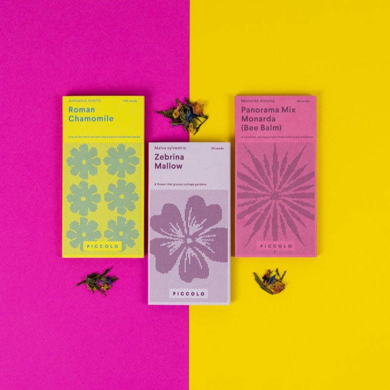 Drei farbenfrohe Piccolo Seeds | Kollektion "Tees mit Blumen" von Piccolo Seeds - gelb, hellviolett und rosa - stehen auf einem gelb-rosa Hintergrund. Jedes hat florale Illustrationen und trägt das Etikett "gentechnikfrei".
