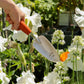 Eine Hand greift nach der Chop Chop | Hochwertige orangefarbene Schaufel 'Tiger Lilly' von Chop Chop Bloom, umgeben von blühenden weißen Schwertlilien und Alliumblüten in einem sonnenbeschienenen Garten.