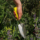 Eine Person hält die Chop Chop Bloom Chop Chop Chop | Hochwertige gelbe Schaufel 'Busy Lizzy' über grüne Pflanzen und lila Blüten in einem Garten.