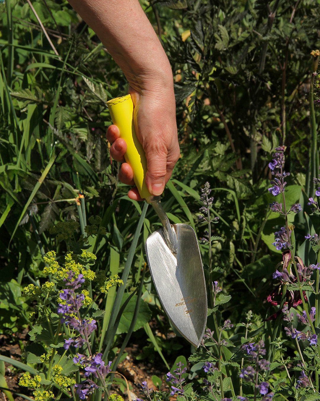 Eine Person hält die Chop Chop Bloom Chop Chop Chop | Hochwertige gelbe Schaufel 'Busy Lizzy' über grüne Pflanzen und lila Blüten in einem Garten.