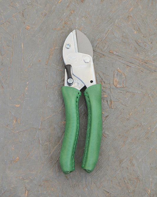 Die Chop Chop Bloom Exklusive Gartenschere 'Dragon Tree' mit silberglänzenden Klingen und grün gepolsterten Ledergriffen bietet präzise Schneidleistung und ruht auf einer strukturierten grauen Holzoberfläche.