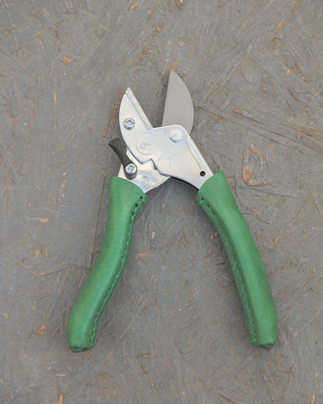 Ein Paar Chop Chop | Exklusive Gartenschere 'Dragon Tree' von Chop Chop Bloom mit grün gepolsterten Griffen ruht auf einer grau strukturierten Oberfläche. Die leicht geöffneten Klingen zeigen die präzise Schneidleistung und robuste Metallmechanik.