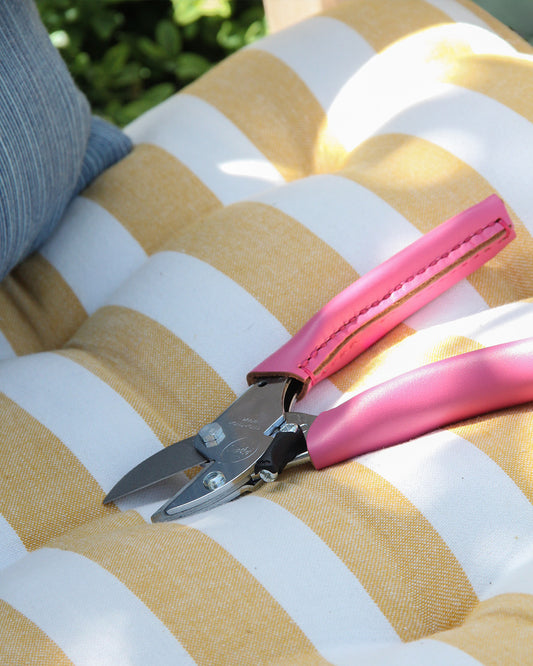 Die hochwertige Gartenschere 'Pink Tulip' von Chop Chop Bloom mit pinken Griffen liegt auf einem gelb-weiß gestreiften Outdoor-Kissen - ein stilvolles Garten-Tool, das die Brustkrebs-Initiative unterstützt.