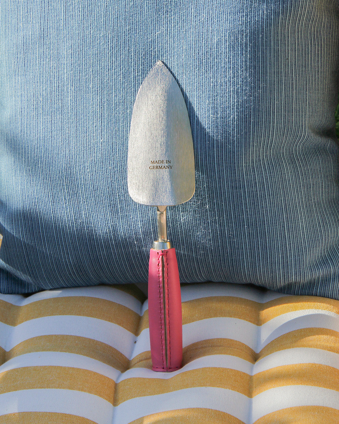 Eine handgefertigte Metall-Schaufel 'Pink Tulip' von Chop Chop Bloom mit pinkem Griff steht auf einem gelb-weiß gestreiften Kissen vor einem blau-weiß gestreiften Polster, "Made in Germany" ist auf der Klinge eingraviert.