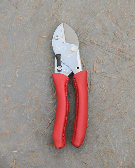 Die Chop Chop Bloom Chop Chop | Exklusive Gartenschere 'Chili Pepper' mit leuchtend roten Griffen ruht auf einer strukturierten grauen Holzoberfläche.