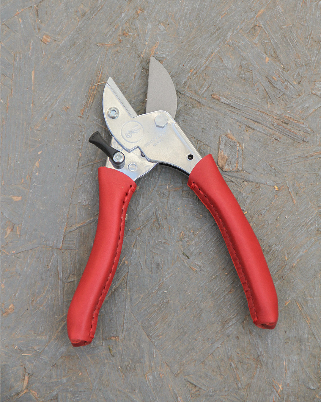 Die Chop Chop Bloom 'Chili Pepper' exklusive rote Gartenschere liegt mit silberner Klinge auf einer strukturierten grauen Oberfläche.