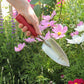 Eine Hand gräbt mit der roten Kelle Chop Chop Bloom 'Chili Pepper' inmitten von blühenden rosa und weißen Blumen in einem üppigen Garten in die Erde.