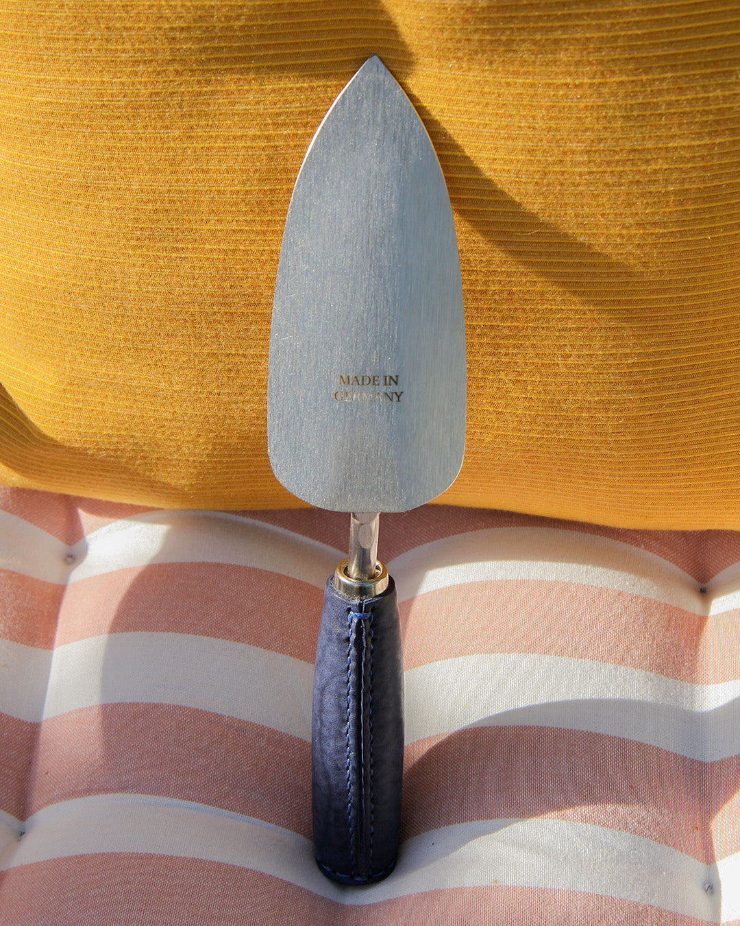 Die handgefertigte, langlebige Schaufel 'Blue Poppy' von Chop Chop Bloom aus Metall mit "MADE IN GERMANY"-Gravur steht aufrecht auf einem gestreiften, gepolsterten Untergrund in Gelb- und Pfirsichtönen.