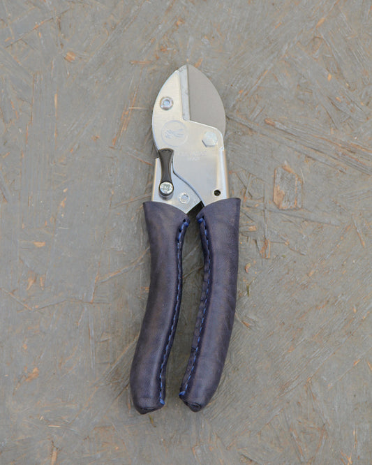 Ein Paar der exklusiven Metall-Gartenschere 'Poppy Blue' von Chop Chop Bloom mit blauen und schwarzen gepolsterten Griffen wird auf einer strukturierten grauen Oberfläche platziert.